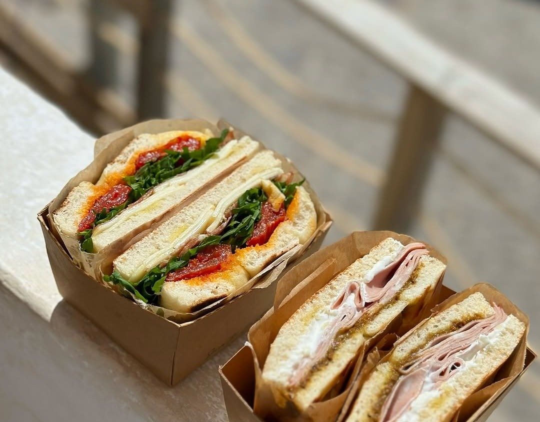 Duas caixas de cartão com sanduíches de focaccia, uma com tomate e rúcula e outra com fiambre, pousadas num muro ao sol