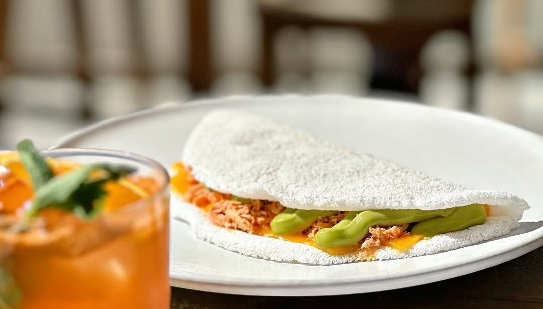 Tapioca recheada com abacate num prato branco, ao lado de um copo com bebida cor de laranja e hortelã